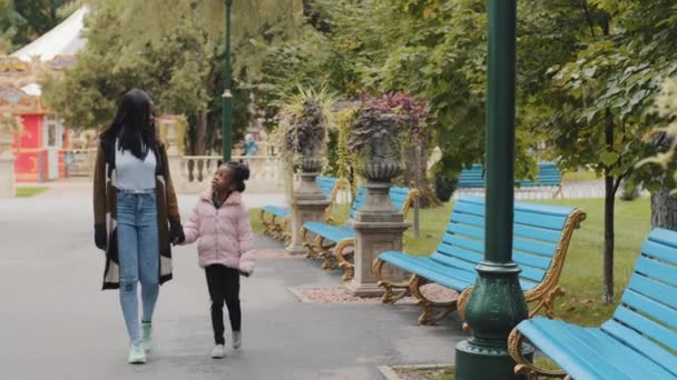 Joven madre caminando con su hija pequeña en el parque de la ciudad en fin de semana alegre alegre familia afroamericana va niño inspirado habla con los padres madre cariñosa sostiene niño pequeño a mano feliz infancia — Vídeo de stock