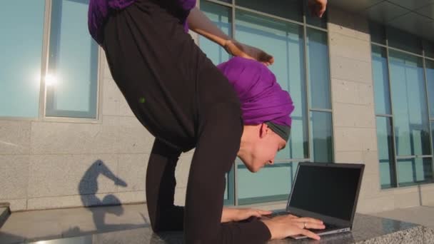 Bewegende beelden moslim meisje student zakenvrouw in hijab werken typen studeren met laptop op straat stad gebouw achtergrond staande in evenwicht yoga asana acrobatische handstand houden benen in de lucht — Stockvideo