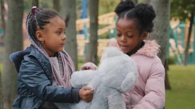 Kızlar kameraya bakıyor. Parktaki bankta oturan iki arkadaş oyuncak ayı kız kardeşlerine okuldan sonra açık havada liseli kızlar oynuyor. Afro-Amerikan çocukları gülümsüyor.