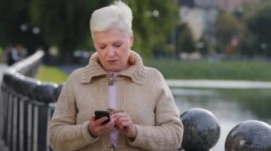 Dışarıda akıllı telefon ekranına bakan hoşnutsuz bir emekli kadın, kötü haber mesajları, spam veya dolandırıcılık mesajlarından memnun değil. Mutsuz, yaşlı bayan mobil uygulama kullanıcısı e-postada phishing linki alıyor