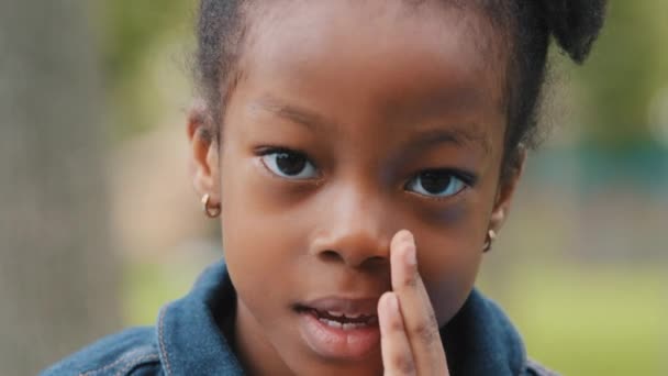 Симпатичный ребенок рассказывает секрет тихо закрывая рот рукой закрывая африканскую маленькую девочку, глядя на камеру ребенок делится секретной информацией на открытом воздухе портрет школьница говорит загадкой снаружи — стоковое видео