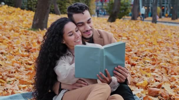 Οι νέοι επικοινωνούν κάθεται σε κίτρινα φύλλα σε εξωτερικούς χώρους ρομαντική ημερομηνία στο πάρκο φθινόπωρο όμορφη σγουρά κορίτσι διαβάσει ισπανόφωνος γενειοφόρος άντρας αγκαλιά σφιχτό δάχτυλο γυναίκα στο βιβλίο αγάπη ζευγάρι χαμογελώντας μαζί — Αρχείο Βίντεο