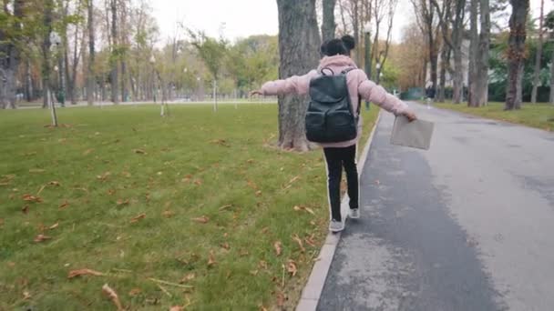 Volver ver colegiala caminar en bordillo niña con mochila negra mantener el equilibrio niño va a la escuela sosteniendo libro en la mano niño desconocido caminando en el parque de la ciudad después de estudiar africano americano alumno jugar solo — Vídeo de stock