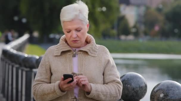 Неймовірна пенсіонерка дивиться на екран смартфона на відкритому повітрі, незадоволена поганими повідомленнями новин, спамом або шахрайськими смс. Нещасливі стресові старша жінка мобільний додаток користувач отримує фішингове посилання в електронній пошті — стокове відео