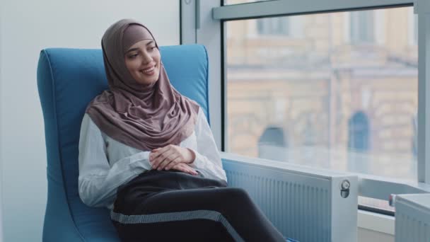 Uśmiechnięta arabska dziewczyna w hidżabie wyglądająca przez okno siedząca w fotelu czekając na wyniki egzaminu lub wywiadu. Kandydat na stanowisko agenta rekrutacyjnego spodziewa się spotkania z pracodawcą w celu zatrudnienia — Wideo stockowe