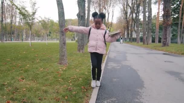 Afro américaine petite fille marche sur bordure bordure équilibrage chemin de l'école dans le parc urbain afro étudiant avec sac à dos profiter passer du temps en plein air en automne enfant tenant livre jouer seul dans la rue de la ville — Video