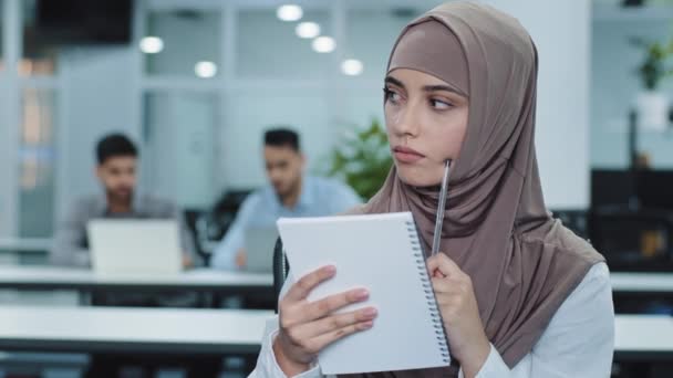 Tankeväckande tusenårig indiansk kvinna i hijab tittar åt sidan, planerar för möte. Pensiva unga arabiska kvinnliga arbetare sitter på kontoret med hjälp av papper anteckningsblock gör anteckningar skapa nya idéer — Stockvideo