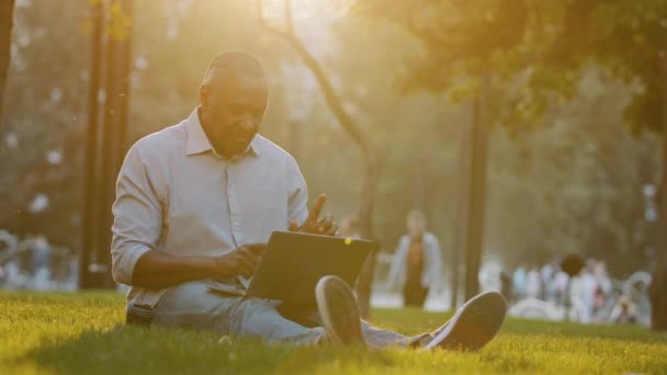 高齢者のフリーランサー作家のジャーナリストやブロガーアフリカ系アメリカ人の男性は、現代のデバイス上でラップトップの無線インターネットを使用して芝生の上に屋外に座って、コンピュータ飲料コーヒーにテキストメッセージを入力します — ストック動画