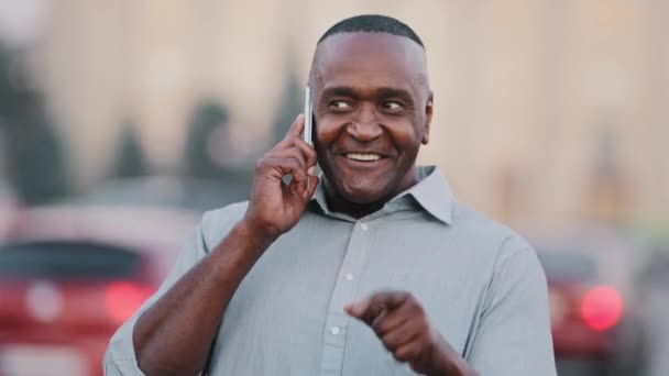 Hombre de negocios afroamericano parado en la calle en el centro de la ciudad sosteniendo un teléfono inteligente, hombre negro profesional divirtiéndose, hablando emocionalmente por teléfono con un amigo o pareja, hombre anciano étnico al aire libre — Vídeos de Stock