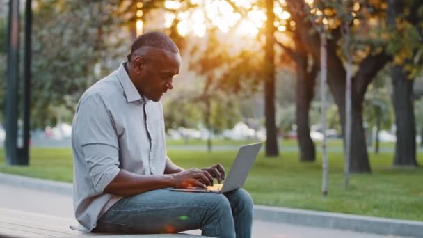 Starszy, afrykański mężczyzna siedzący na zewnątrz, przeglądający bezprzewodowy Internet na nowoczesnym urządzeniu, wpisujący SMS-y na laptopie pracującym na komputerze, dorosły freelancer konsultuje się z klientem online, koncepcja technologii — Wideo stockowe
