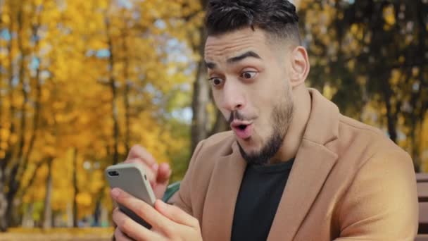 Podekscytowany Hiszpan czyta dobre wieści na smartfonie siedzi na ławce w jesiennym parku młody człowiek szczęśliwie zaskoczony otrzymaniem wiadomości przeszczęśliwy mężczyzna zwycięzca świętuje zwycięstwo trzymając telefon na zewnątrz — Wideo stockowe