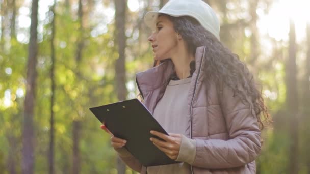 Milenial mujer técnica ecologista mirando hacia arriba en las copas de los árboles, Joven mujer india en hardhat con portapapeles tomando medidas de comprobación de los árboles. Ingeniero forestal en el parque. Supervisar el santuario de vida silvestre — Vídeo de stock