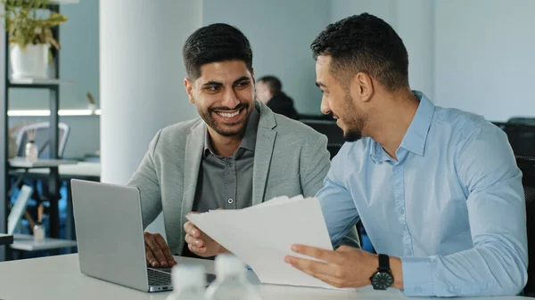 Twee succesvolle Arabische mannelijke zakenmannen in functie met papieren documenten. Diverse lachende jonge collega 's Indiase medewerkers kijken naar laptop scherm ontvangen goed nieuws, bespreken goede deal — Stockfoto