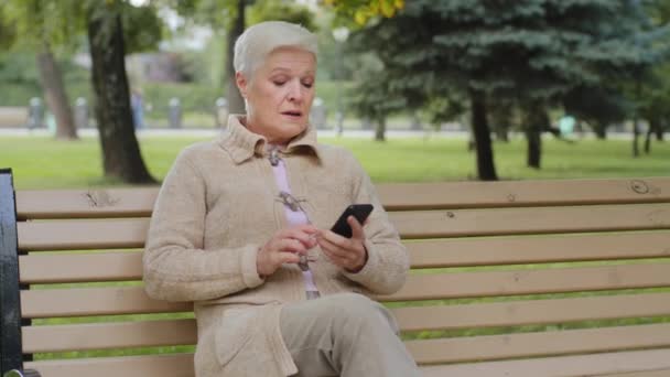 Attraente donna anziana dai capelli grigi in età pensionabile seduta sulla panchina del parco che fa videochiamate, guardando la fotocamera dello smartphone. Nonna in pensione utilizza gadget moderno internet parlando sul telefono cellulare all'aperto — Video Stock