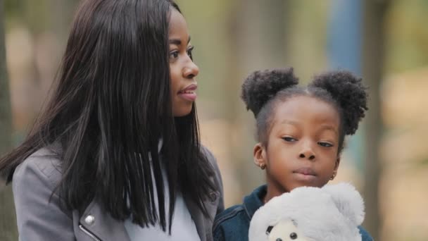 Закройте счастливую африканскую американскую семью улыбаясь в парке маленькая девочка разговаривает с женщиной ребенок держит плюшевый медведь портрет мать и дочь глядя в сторону ребенка весело общаться с молодой мамой на открытом воздухе — стоковое видео