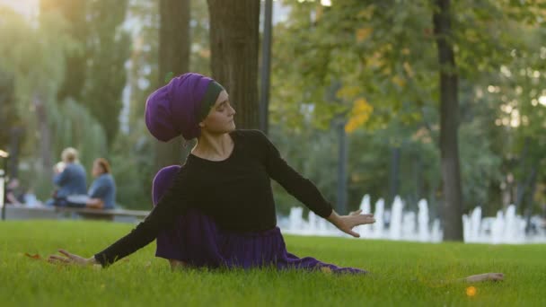 Muzułmańska dziewczyna w hidżabie islamska kobieta Sportswoman jogi ćwiczy jogę w parku na zielonym trawniku siedzi w sznurku z jedną nogą zgięcia rozciąga się robi uchwyt za plecami rozciągając elastyczność — Wideo stockowe