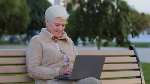 Wiek kobieta siedzi na ławce w parku miejskim za pomocą Internetu surfingu komputerowego, uśmiechnięta starsza kobieta w wieku emerytalnym pracuje w laptopie czytanie wiadomości online, starsza pani smsowanie wiadomości na zewnątrz komputera — Wideo stockowe