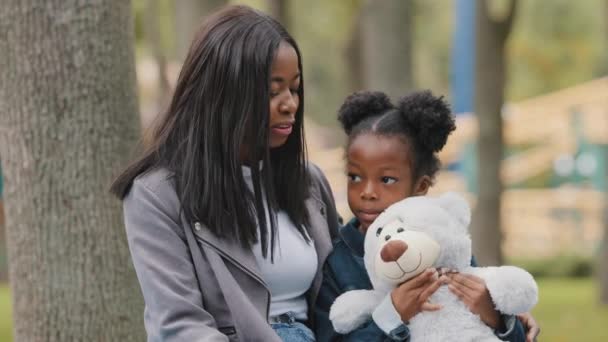 Lánya fiatal anya ül padon a városi parkban kis aranyos lány kezében játék medve kezében afro-amerikai anya kommunikál és baba szabadban anyu ölelés gyermek család együtt boldog időt töltenek