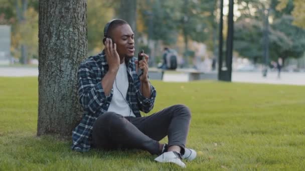 Afroamerikansk ung man kille student man bär rutig skjorta sitter på gräs i parken på grön gräsmatta lyssna musik på hörlurar online ljud ljud app sjunga i telefon som mikrofon njuter av sång — Stockvideo