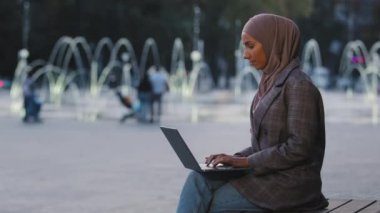 Şehirli fıskiyelerde çalışan, tesettürlü genç müslüman kız tesettüre bakıp mutlu gülümseyen dizüstü bilgisayar uygulaması kullanıyor. Kablosuz bilgisayarı olan Arap kadın.