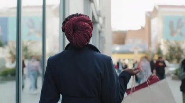 Arkaya bakan Afro-Amerikalı alışveriş meraklısı tüketici alışveriş torbalarıyla alışveriş yaptıktan sonra şehrin yakınındaki caddede yürüyen kendinden emin ve kameraya bakan bir kadın.