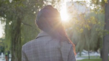Kıvırcık, uzun siyah saçlı, kıvırcık saçlı, güzel, şık, çekici bir kadın parkta yürüyor gün batımında güneş ışığı arka planında kameraya bakarak gülümsüyor.