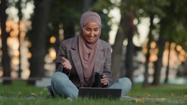 Yeşil çimen parkındaki tesettürlü neşeli müslüman kız video konferansı için dizüstü bilgisayarını kullanıyor. İnternet üzerinden merhaba diyor, internet kamerasıyla kablosuz bilgisayarla sohbet ediyor.