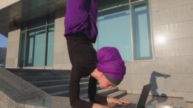 Mor tesettürlü güçlü müslüman iş kadını, İslamcı kız kullanıcı alışılmadık poz veriyor asana denge yoga akrobatiği, kablosuz bilgisayar kullanarak laptopla çalışıyor.