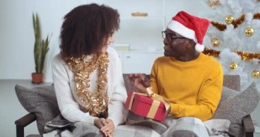 Kıvırcık saçlı kız arkadaş eşi kız kardeşi kocasına siyah erkek Afrikalı Amerikalı çocuğa hediye paketi veriyor. Kırmızı ambalajlı Noel hediyesi. Yeni yıl tatilini evde koltukta oturarak kutluyor.