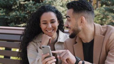 Güz parkında çekici bir kızla sakallı adam neşeli bir çiftin yanında oturmuş akıllı telefon ekranına bakıyor genç adam aktif olarak kız arkadaşlarıyla iletişim kuruyor online alışverişi tartışıyorlar.