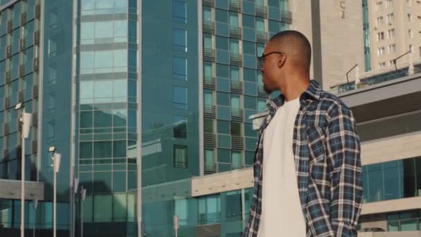 Élégant afro-américain gars modèle étudiant réussi homme d'affaires touriste homme à la mode lunettes de soleil et chemise à carreaux se tient dans le fond du bâtiment de la ville tournant souriant à la caméra bras croisés — Video