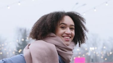 Yakın plan güzel, melez, Afro-Amerikan kadın bayan bayan bayan öğrenci gülümsemesi takıyor soğuk mevsimde şehir sokaklarında eşarp stantları kışın gökyüzüne bakıp gülümsüyor ve kameraya bakıp gülüyor.