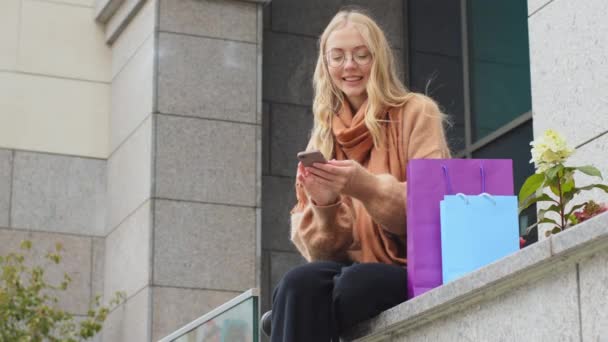 Femme shopper assis à l'extérieur près du bâtiment avec des sacs colorés se réjouit vente discount noir vendredi attrayant fille souriant achats en ligne en utilisant téléphone heureuse femme messagerie tenant le téléphone dans la main — Video