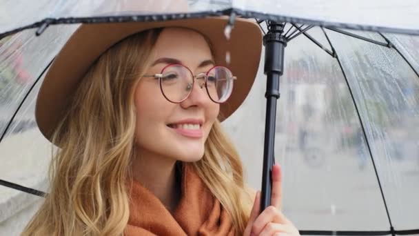 Attraktiv ung flicka står på gatan under paraply närbild kaukasiska kvinna ler med vita tänder porträtt glad bekymmerslös dam njuter av regn utomhus europeiska blond kvinna med hatt och glasögon — Stockvideo