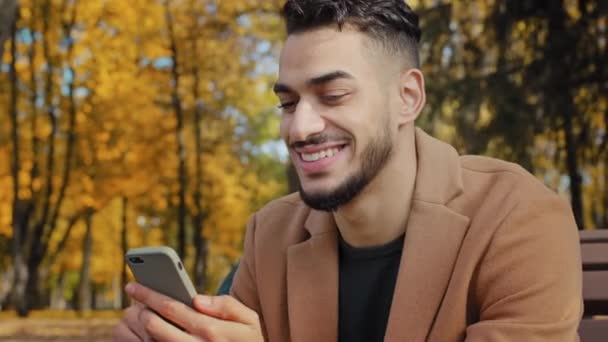Gros plan jeune hispanique gars assis sur le banc en automne parc barbu homme tenant téléphone mobile rend achats en ligne sourire heureux en utilisant l'application regardant dans smartphone en plein air nouvelles de navigation sur l'appareil — Video
