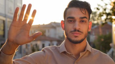 Şehirde erkek portresi. Genç yetişkin, ciddi, sakallı iş adamı. Gömlekli patron. Gün batımındaki güneş ışınlarının arka planında el ele tutuşan kameraya bakıyor.