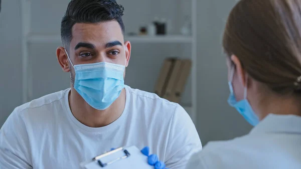 Arab férfi spanyol férfi beteg beteg férfi visel orvosi védő maszkot az arcon beszél tünetei egészségügyi problémák felismerhetetlen orvos nő nővér leírja jegyzetek, konzultáció a klinikán — Stock Fotó