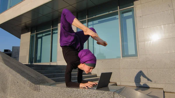 Silný akrobat obchodní žena legrační muslim dívka v hidžábu stojí na rukou v akrobatické józe póza asana rovnováhu handstand ve městě venku psaní notebook pracuje dálkově on-line udržet nohy ve vzduchu — Stock fotografie