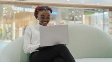 Mutlu gülümseyen Afro-Amerikan öğrenci iş kadını serbest çalışan ofis alışveriş merkezinde oturmuş bilgisayardaki internetteki wi-fi uygulamasını yazarak sohbet ediyor.