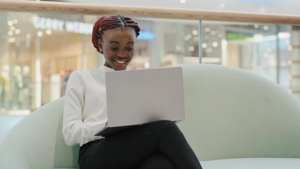 Feliz chica sonriente afroamericana estudiante de negocios mujer freelancer sentado en el sofá en el centro comercial de la oficina de chat escribiendo en el ordenador portátil aplicación en línea wi-fi red de trabajo remoto e-learning — Vídeos de Stock