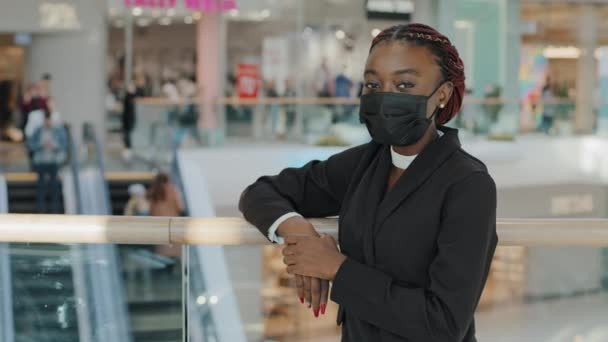 Afro-Amerikan Afro kadın tüketici portresi alışveriş merkezinde durmuş salgın zamanında dükkan yürüyen merdivenin yanında poz veren kameralara bakarken siyah tıbbi maske takıyor. — Stok video