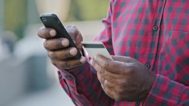 Afrikalı Amerikalı adamın cep telefonuyla kredi kartıyla alışveriş yaptığı yakın çekim. Çift ırklı erkek alıcı müşteri siparişi akıllı telefondan güvenli ödeme, açık havada internet alımı
