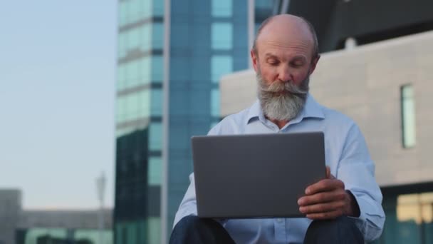 Szczęśliwy starszy europejski senior korzystający z laptopa outdoor, dokonujący płatności, dokonujący zakupów online. Atrakcyjny dziadek z brodą patrzy na ekran komputera, wysyła lub odbiera wiadomości e-mail w posłańcu — Wideo stockowe