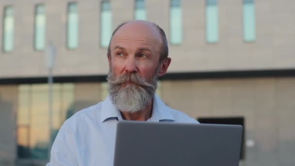 Pensiv äldre man med grått skägg kreativ professionell arbetar på laptop utomhus tittar åt sidan, funderar idé. Gammal manlig pensionär tillbringar fritid på datorn fungerar på distans skickar e-post — Stockvideo