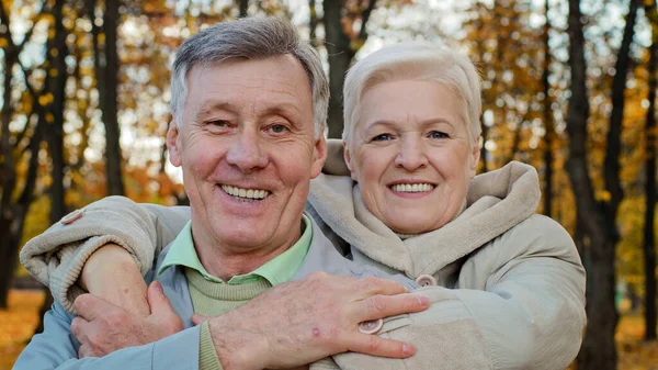 Dospělá stará žena objetí milovaný manžel podle ramen starší ženatý pár objímání v podzimním parku prarodiče s úsměvem na kameru pak každý jiný těšit komunikace venkovní šťastný manželství koncept — Stock fotografie