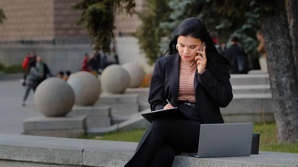 年轻的女商人在户外用电脑上网，用手机聊天，找出工作时间；女人在纸上写数据；女人坐在街上做着多项任务，让漂亮的西班牙女人惊慌失措 — 图库照片