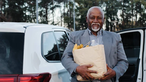 ハンサムな古いアフリカ系アメリカ人男性の手で果物店で買った食品を保持笑みを浮かべて幸せな屈託のない年金受給者男性のクローズアップは、前の白い車の中でスタンド高齢者のカメラを見て灰色の髪のひげ — ストック写真