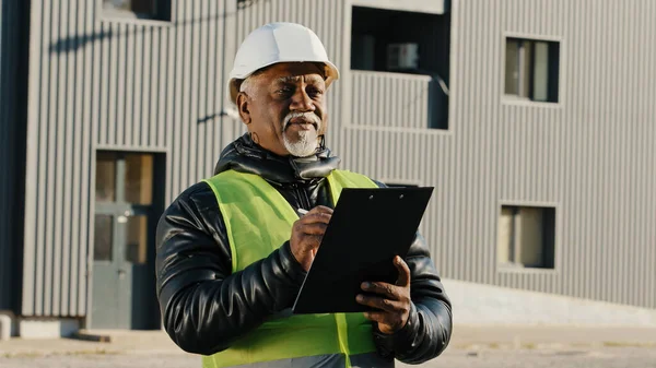 Anciano afroamericano gerente de construcción hombre evalúa edificio viejo capataz de pie en la calle en hardhat protector escribe datos a tableta ingeniero constructor piensa en primer plano plan de trabajo al aire libre —  Fotos de Stock