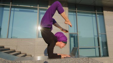 Başörtüsü içinde komik bir kız dizüstü bilgisayarında yazı yazan kullanıcı kadın baş aşağı yoga egzersizi yapan asana şehir caddesinde bilgisayarla uzaktan öğrenim görüyor.
