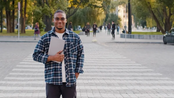 Bem sucedido jovem afro-americano homem de negócios cara africano estudante em óculos de sol fica na rua da cidade andando travessia estrada trânsito carro pedestre cruzamento fundo segurando laptop sorrindo indo caminhadas — Fotografia de Stock